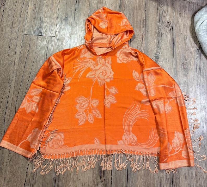 May include: An orange hooded scarf with a floral rose pattern. The scarf has a fringe along the bottom edge and is made of a soft, flowing material. The hood is attached to the scarf, creating a cozy and stylish accessory.