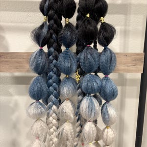 May include: Braided hair extensions in shades of black, blue, and white, adorned with gold-colored accents. The extensions are arranged in a vertical display, showcasing the color gradient and textures of the hair pieces.