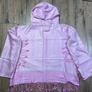 May include: A light pink hooded top with long sleeves and fringe detailing. The top has a horizontal stripe design and is made of a silky material. The hood is attached to the neckline. The garment is displayed on a wooden surface.