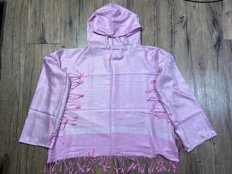 May include: A light pink hooded top with long sleeves and fringe detailing. The top has a horizontal stripe design and is made of a silky material. The hood is attached to the neckline. The garment is displayed on a wooden surface.