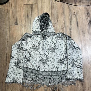 May include: A hooded shawl with a white base and a black paisley pattern. The shawl has a fringe along the bottom edge and a black and white patterned hood. The paisley design is repeated throughout the fabric.