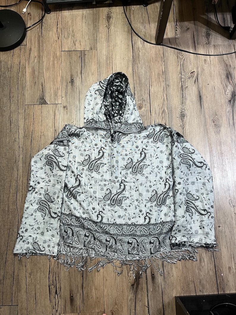 May include: A hooded shawl with a white base and a black paisley pattern. The shawl has a fringe along the bottom edge and a black and white patterned hood. The paisley design is repeated throughout the fabric.