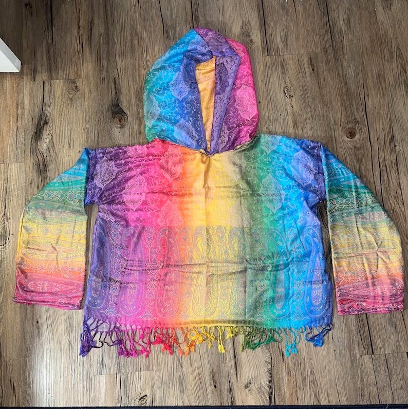 May include: A hooded poncho in a vibrant rainbow of colors, including pink, yellow, green, and blue. The poncho features a paisley pattern and fringe along the bottom. The sleeves are wide and flowing.