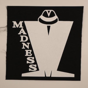 May include: A black square patch with a white graphic of a figure wearing a hat, with the word "MADNESS" in white capital letters on the left side. The patch has a textured surface.