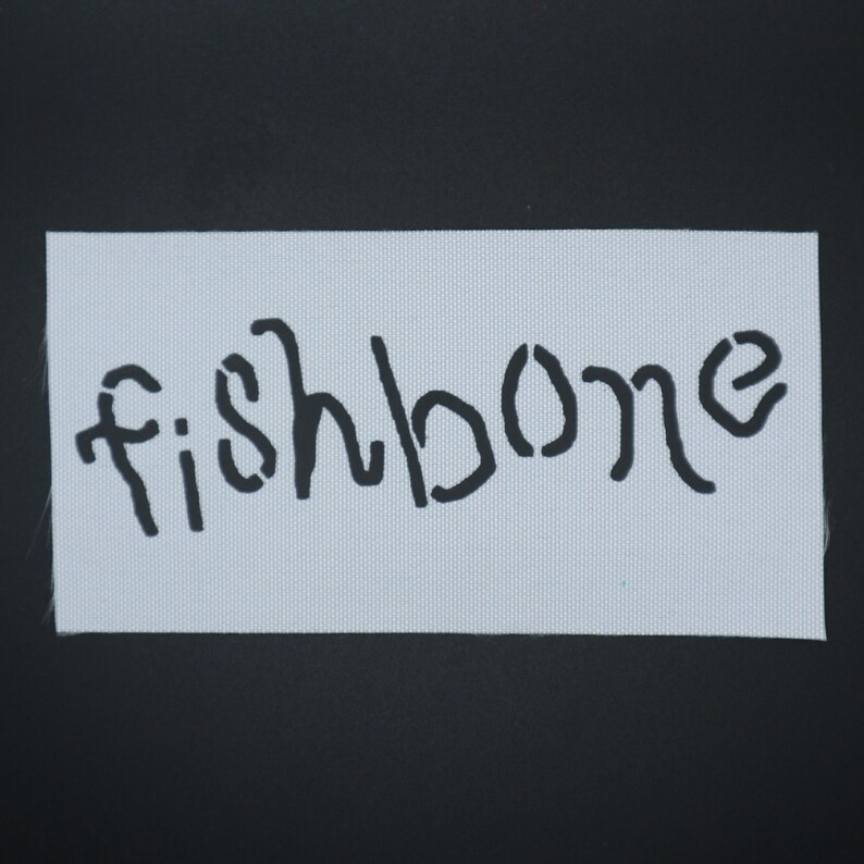 Op de afbeelding: Een rechthoekige witte stoffen patch met het woord "fishbone" in zwart geschilderd. De stof heeft een subtiel, getextureerd patroon. De achtergrond is effen zwart.
