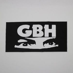 May include: Black rectangular patch with the white letters "GBH" above a white stylized eye design. The patch is on a white background.