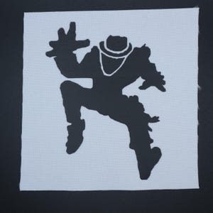 May include: A black silhouette of a figure in a hat and necklace, in a dynamic pose, is centered on a square, light blue fabric. The figure's arms are outstretched, and one leg is bent, suggesting movement. The fabric has a textured appearance.