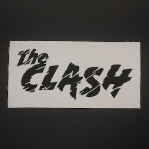 May include: A rectangular white patch with the words "the CLASH" in bold, black, distressed lettering. The patch has a woven texture and is set against a black background. The design is a classic punk rock aesthetic.