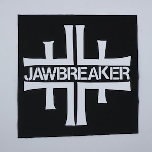 May include: A square black fabric patch featuring a white graphic design of a stylized cross shape with the word JAWBREAKER printed in white capital letters across the center of the design. The patch has a visible woven texture throughout.