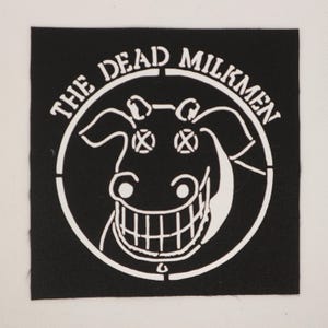 May include: Black square patch featuring a white graphic of a grinning cow's head inside a circle, with the text "THE DEAD MILKMEN" around the edge. The design is a stencil style.