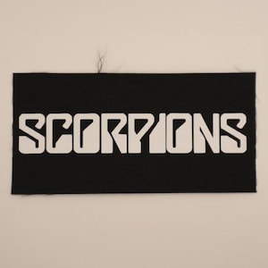 May include: A rectangular black fabric patch featuring the word SCORPIONS printed in a bold, white, stylized sans-serif font. The patch has a visible woven texture and clean, straight edges, designed for application onto clothing or other textile items.