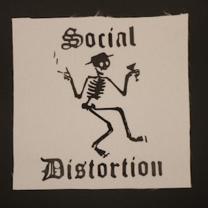 May include: A white square patch with the words "Social Distortion" in gothic lettering. A black skeleton figure is depicted wearing a hat, holding a cocktail, and smoking a cigarette.