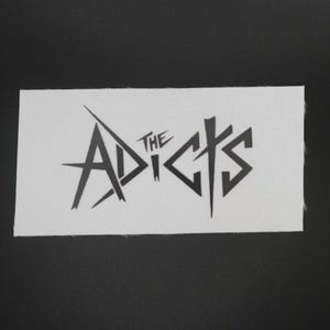 May include: A white rectangular patch with the black text "THE ADICTS" in a stylized, angular font. The patch is on a black surface.