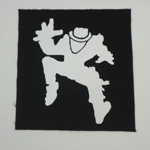 May include: Square black patch with a white silhouette of a person in a dynamic pose, wearing a hat and a scarf. The figure is mid-jump with one arm raised. The patch is likely for clothing or accessories.