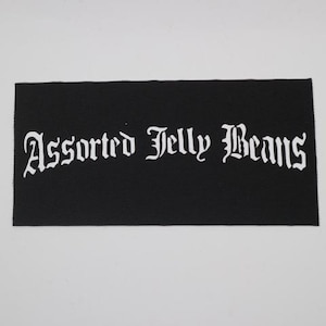 May include: A black rectangular patch with the words "Assorted Jelly Beans" in white gothic lettering. The patch is made of a textured fabric and is set against a white background.