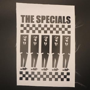 May include: A white fabric patch featuring the band name "THE SPECIALS" above a black and white checkered pattern. Below the pattern are five stylized figures in suits and sunglasses, also in black and white.