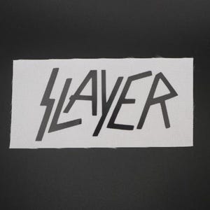 May include: A rectangular white patch with the word "SLAYER" in black, stylized lettering. The "S" is a lightning bolt. The patch is made of a textured fabric, and is set against a black background.