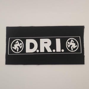 May include: A rectangular black fabric patch featuring a white printed design. The graphic includes the text D.R.I. in the center, flanked on both sides by a circular icon depicting a stylized running figure in white on a black background.