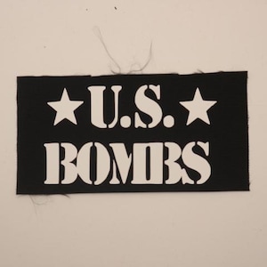 May include: Black rectangular patch with white text and stars. The text reads "U.S. BOMBS" in a bold, stencil-style font, with a star on each side. The patch is likely for clothing or accessories.
