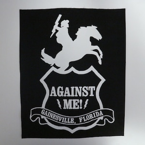 May include: Black patch featuring a white graphic of a rider on a rearing horse, holding a lightning bolt. The text "AGAINST ME!" is above "GAINESVILLE, FLORIDA" on a banner. A shield shape frames the text and rider.