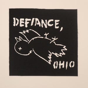 May include: A square black fabric patch featuring a white screen-printed graphic of a bird with an X over its eye and feet pointing upward. The text DEFIANCE, is printed above the bird and the word OHIO is printed below the bird in white.