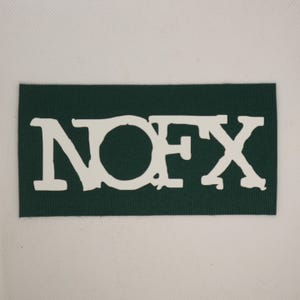 Peut inclure: Un &eacute;cusson rectangulaire vert fonc&eacute; avec le texte blanc "NOFX". Les lettres ont un aspect vieilli et textur&eacute;. L'&eacute;cusson est pos&eacute; sur un fond blanc.