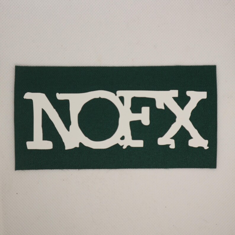 Peut inclure: Un &eacute;cusson rectangulaire vert fonc&eacute; avec le texte blanc "NOFX". Les lettres ont un aspect vieilli et textur&eacute;. L'&eacute;cusson est pos&eacute; sur un fond blanc.