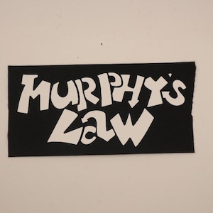 May include: A black rectangular patch with the words "MURPHY'S LAW" in large, white, stylized lettering. The patch has a textured appearance and a raw edge.
