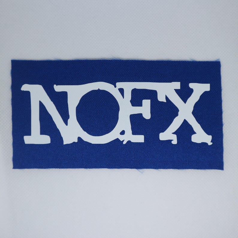 Peut inclure: Un &eacute;cusson rectangulaire bleu avec le texte blanc "NOFX". Les lettres ont un aspect vieilli et textur&eacute;. L'&eacute;cusson est fait d'un tissu tiss&eacute; et est pos&eacute; sur un fond blanc.