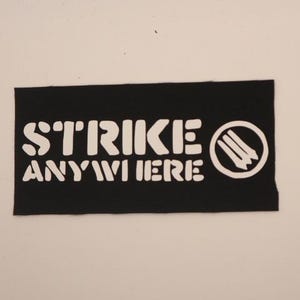 May include: A rectangular black fabric patch featuring the white text STRIKE ANYWHERE in a bold, rounded font. To the right of the text is a white circular emblem containing three vertical, pointed shapes resembling matchsticks or stylized lines.