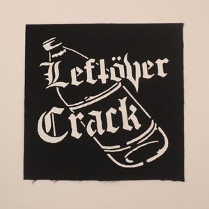 May include: Black square patch with white text and a bottle illustration. The text reads "Leftöver Crack" in a gothic font, with the bottle design overlaying the words. The patch is likely for clothing or accessories.