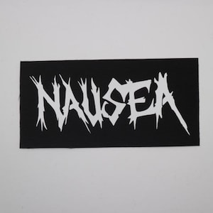 May include: A black rectangular patch with the word "NAUSEA" in bold, white, stylized lettering. The letters have sharp, jagged edges, giving a distressed appearance. The patch is likely for clothing or accessories.