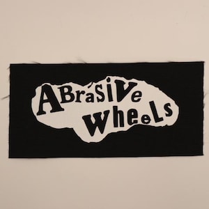 May include: A black rectangular patch with the words "Abrasive Wheels" in white, stylized lettering. The text is set against a white, irregular shape. The patch has a textured surface.