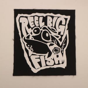 May include: Black square patch with white text and a cartoon fish design. The text reads "REEL BIG FISH" in a bold, stylized font. The fish has large eyes and is facing to the right. The patch is likely for a jacket or bag.