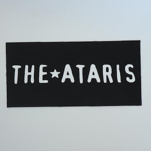May include: Black rectangular patch with the text "THE STAR ATARIS" in white, sans-serif font. The star is between the words "THE" and "ATARIS". The patch has a textured surface and is set against a white background.