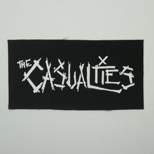 May include: Black rectangular patch with the white text "THE CASUALTIES" in a stylized, graffiti-like font. The patch is likely made of fabric and is suitable for sewing or ironing onto clothing or accessories.