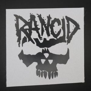 May include: Square white patch with a black graphic of the word "RANCID" above a skull with a radiation symbol. The patch has a woven texture and is likely for clothing or accessories.
