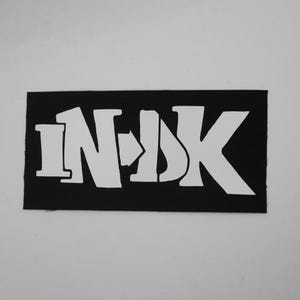 May include: A black rectangular patch with the white letters "INK" in a stylized font. The patch has a textured surface and is set against a white background. The letters are bold and modern.