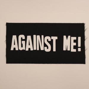 May include: A black rectangular patch with the words "AGAINST ME!" in bold white capital letters. The patch has a textured surface and frayed edges, set against a plain white background.