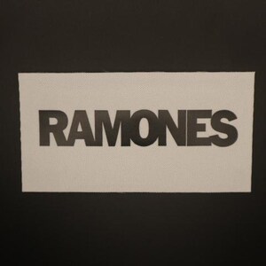 May include: A rectangular patch with the word "RAMONES" in bold, black capital letters. The patch has a white background with a textured appearance, set against a black backdrop. The design is simple and graphic.