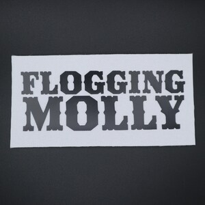 May include: A white rectangular patch with the words "FLOGGING MOLLY" in bold, black, block letters. The patch has a textured surface and is set against a dark gray background.