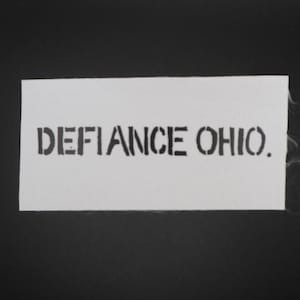 May include: A white rectangular patch with the words "DEFIANCE OHIO." stenciled in black, centered on the patch. The patch is set against a black background.