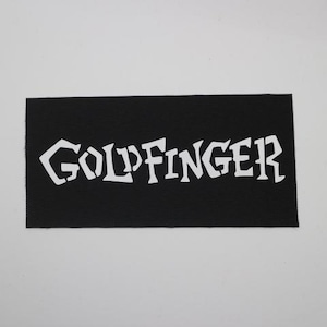 May include: A black rectangular patch with the word "GOLDFINGER" in white, stylized, block letters. The patch has a textured surface and is set against a white background.