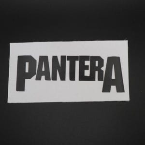 May include: A white rectangular patch with the word "PANTERA" printed in bold, black capital letters. The patch is set against a black background.