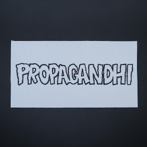 May include: A rectangular white patch with the word "PROPAGANDHI" in black, outlined, bubble letters. The patch has a textured surface and is set against a dark background.