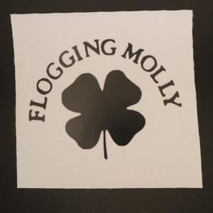 May include: A white square patch with the words "Flogging Molly" arched above a black four-leaf clover. The text and clover are centered on the patch, which is set against a black background.