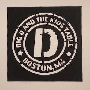 May include: White and black circular patch with the text "Big D and the Kids Table Boston, MA" and a large white letter "D" in the center.