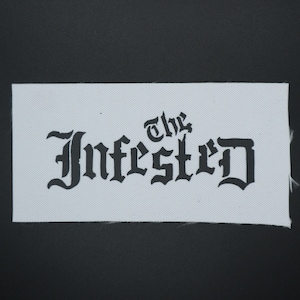May include: A white rectangular fabric patch with the words "The Infested" printed in black gothic lettering. The patch is set against a black background, highlighting the stark contrast between the text and the fabric.