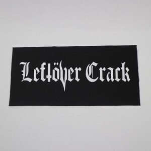 May include: Black rectangular patch with the words "Leftöver Crack" in white gothic lettering. The patch is made of a woven fabric and is likely designed to be sewn onto clothing or accessories.