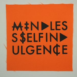May include: An orange fabric square with black text. The text includes the words "MINDLESS," "SELF," and "URGENCE," along with symbols like a dollar sign, a play button, and a cent symbol. The fabric has a textured appearance.